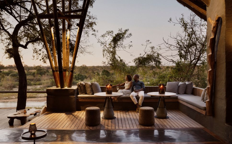 Couple relaxing with drinks on a candlelit outdoor lounge deck at Singita Boulders Lodge, overlooking the serene South African bush at sunset.