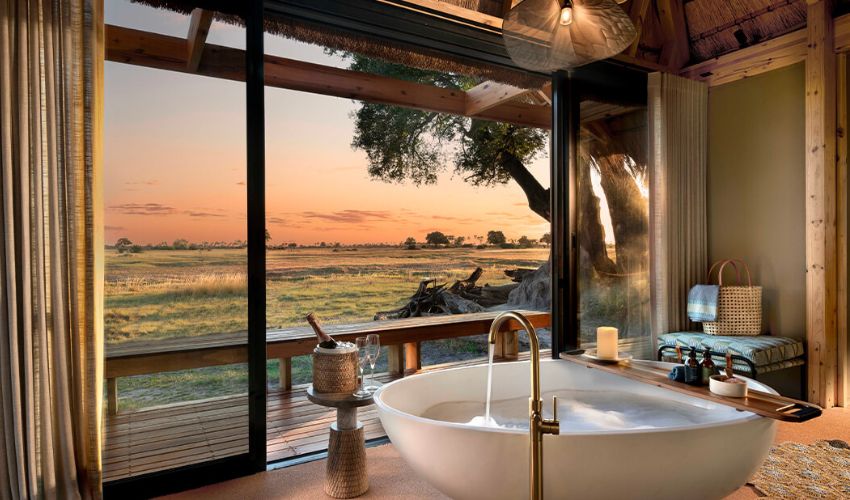 A luxurious freestanding bathtub in an open-air suite at Tawana Camp, overlooking the vast Okavango Delta at sunset. A glass of champagne and a candlelit ambiance create the perfect romantic retreat in Botswana’s wilderness.
