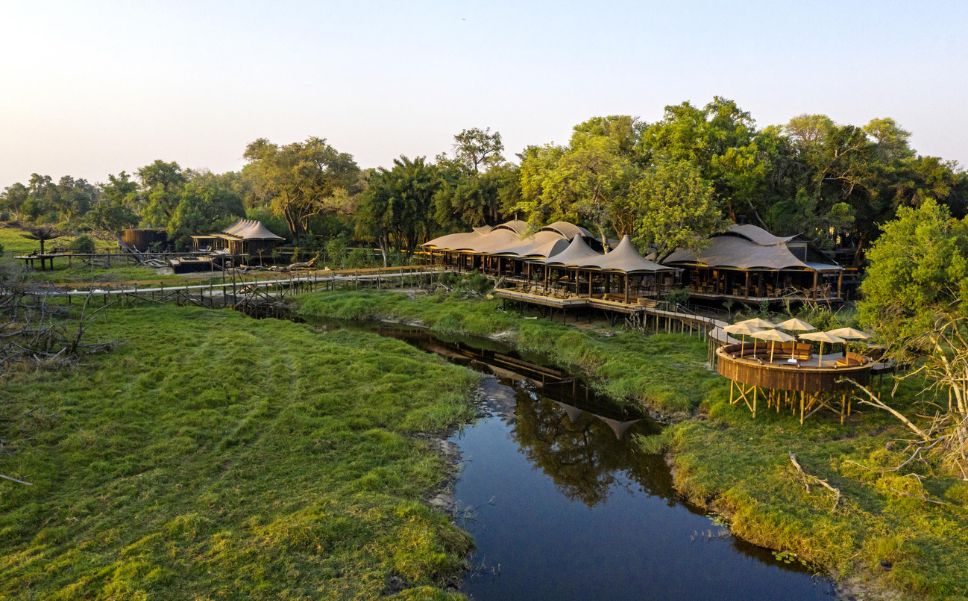 Aerial view of a Xigera Safari Lodge nestled along a channel in Botswana's Okavango Delta, surrounded by lush greenery and elevated wooden walkways.