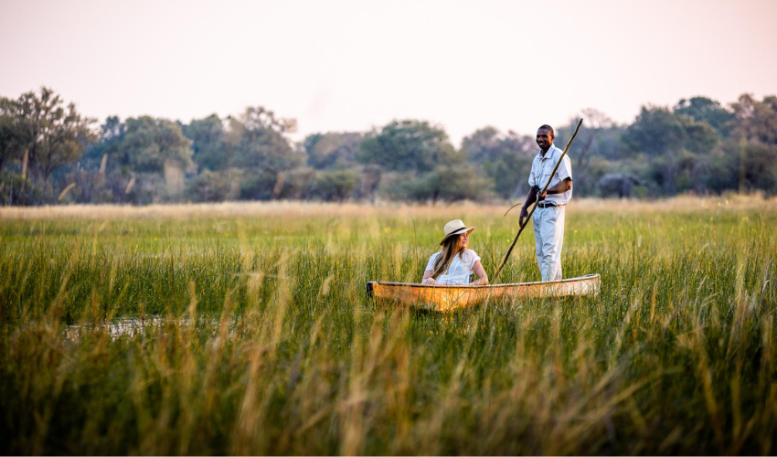Guest and guide exploring the Okavango Delta in a traditional mokoro canoe surrounded by tall grasses and golden light.